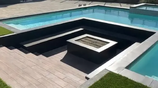 The image shows a pool, with inset stairs and fire pit. The pool's design is modern and angular, featuring a rectangular shape and integrated seating areas.